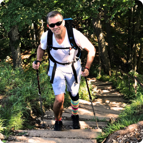 Foto von einem Mann mit einem bunt geringelten Strump am linken Bein, der mit Nordic Walking Sticks im Wald eine Treppe hochläuft